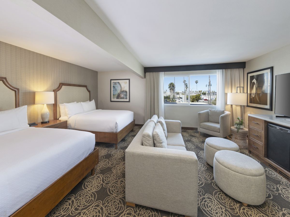 A modern hotel suite with two beds, lounge chairs, lamps, a TV, and a window view of palm trees.