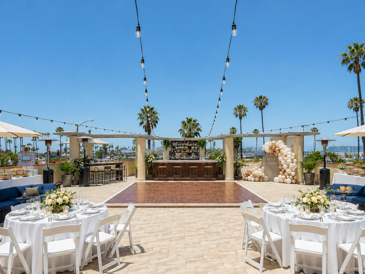 Outdoor wedding/event patio with tables, string lights, umbrellas, a bar, and palm trees under a bright blue sky.