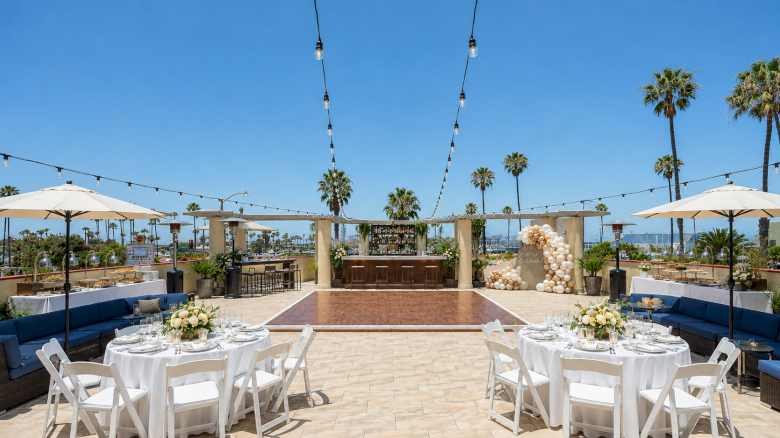 Outdoor wedding/event patio with tables, string lights, umbrellas, a bar, and palm trees under a bright blue sky.