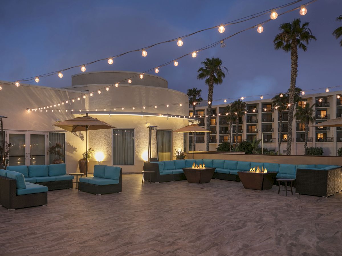 A chic rooftop lounge with blue sectional seating, string lights, palm trees, and a curved white cabana at dusk, inviting relaxation.