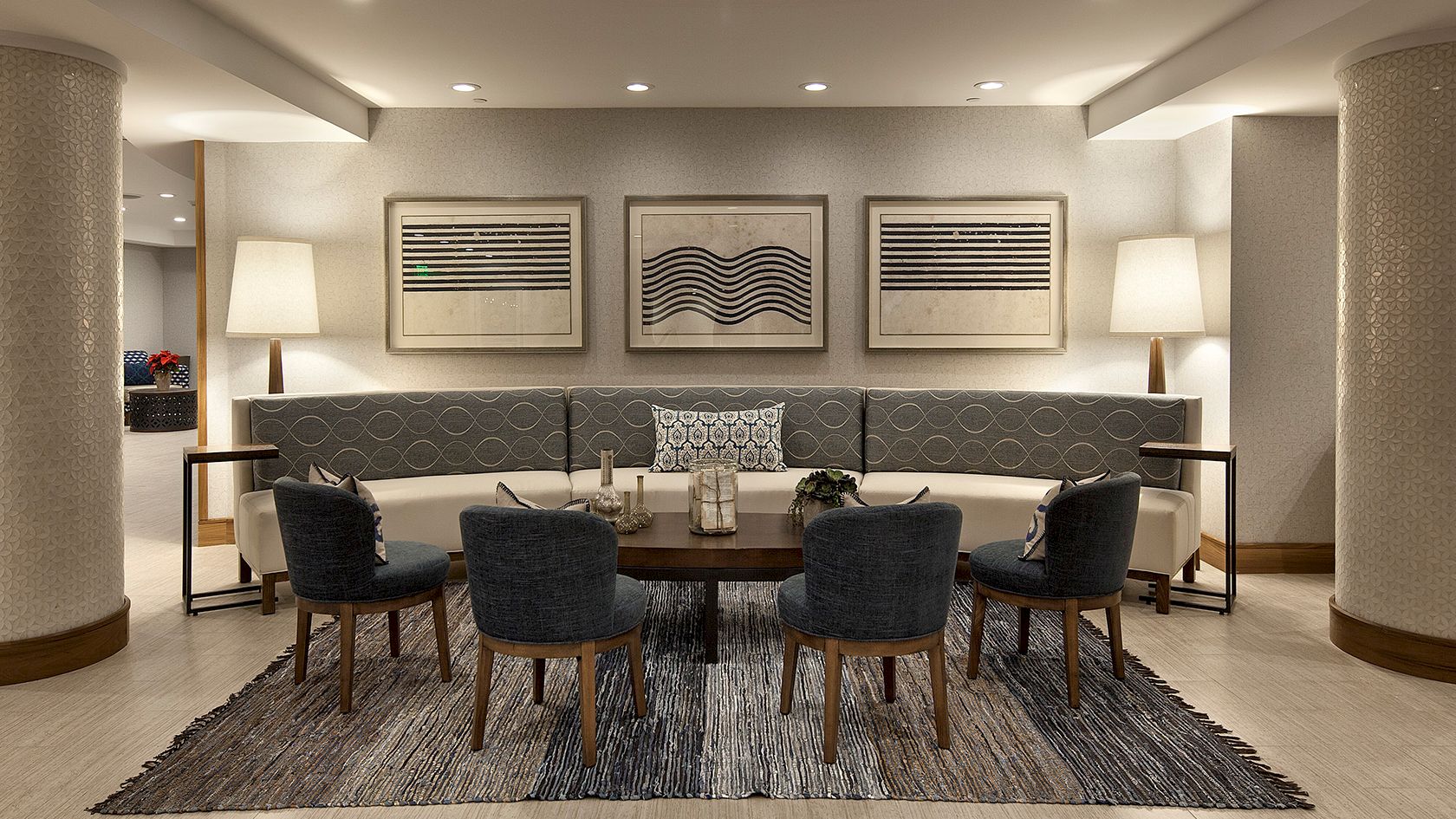A modern hotel lounge with a circular seating area, gray chairs, a dark rug, framed wall art, and tall lamps on either side. Stop.