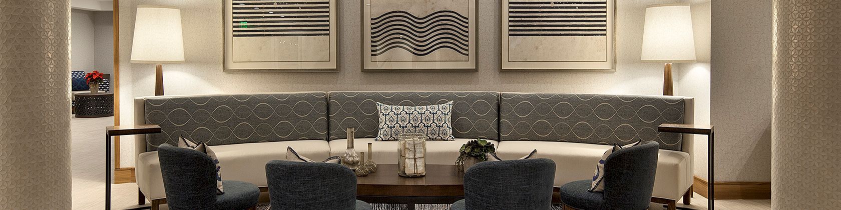 A modern hotel lounge with a circular seating area, gray chairs, a dark rug, framed wall art, and tall lamps on either side. Stop.