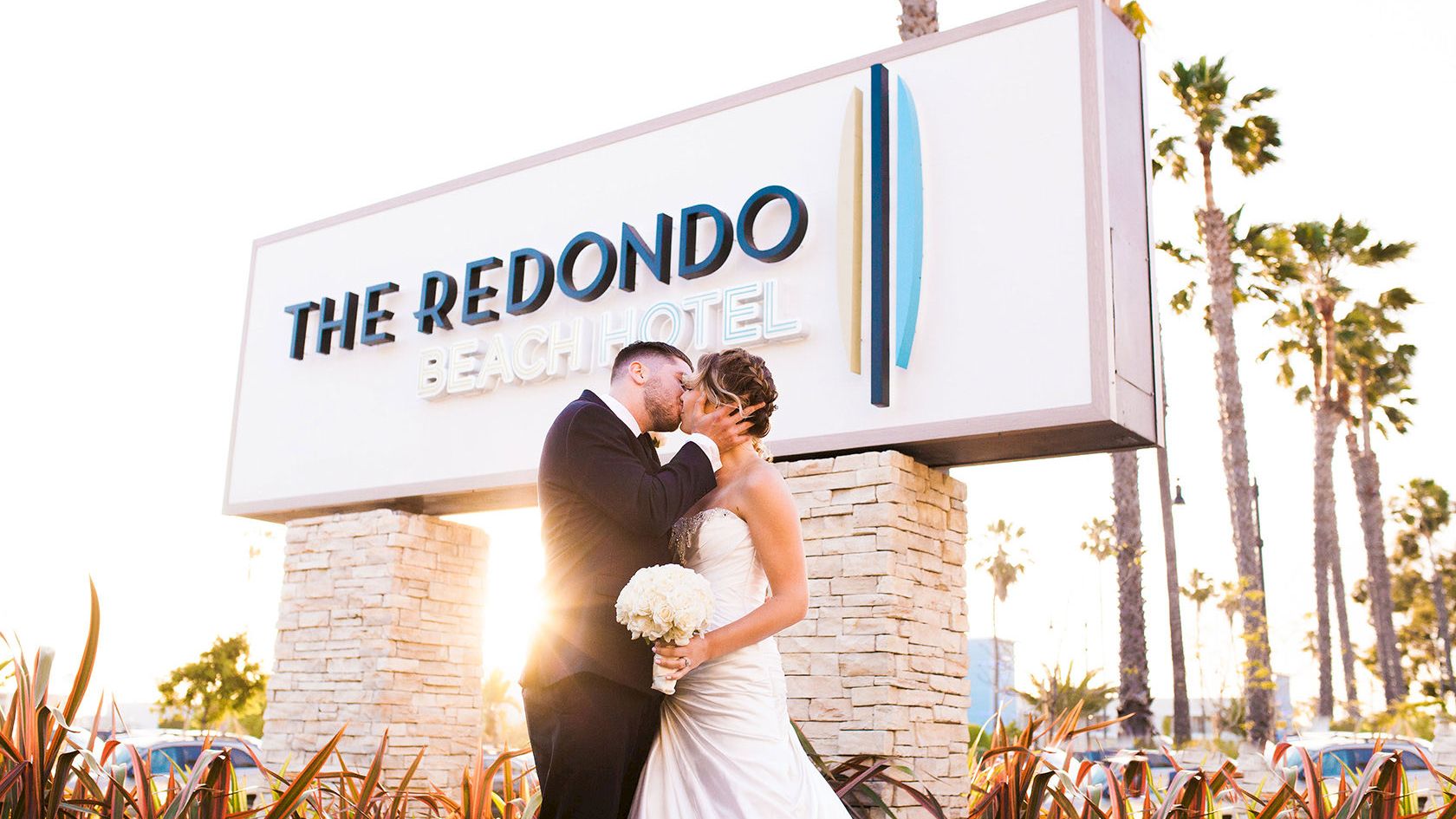 Two newlyweds kiss in front of The Redondo Beach Hotel sign, bride in a white gown holding a bouquet, sunny palm-lined backdrop.