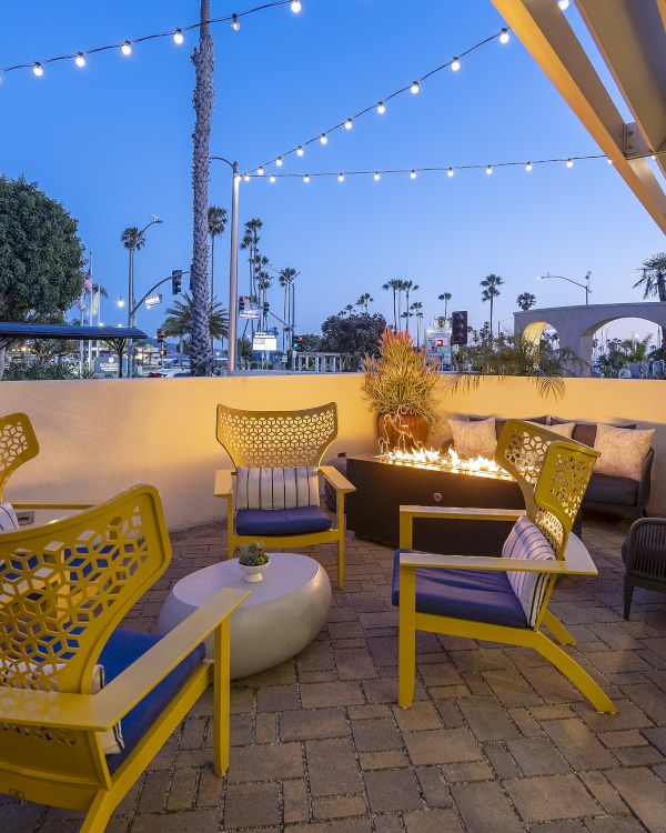An outdoor patio with yellow chairs, a glowing fire pit, string lights, and palm trees at dusk by a waterfront walkway.