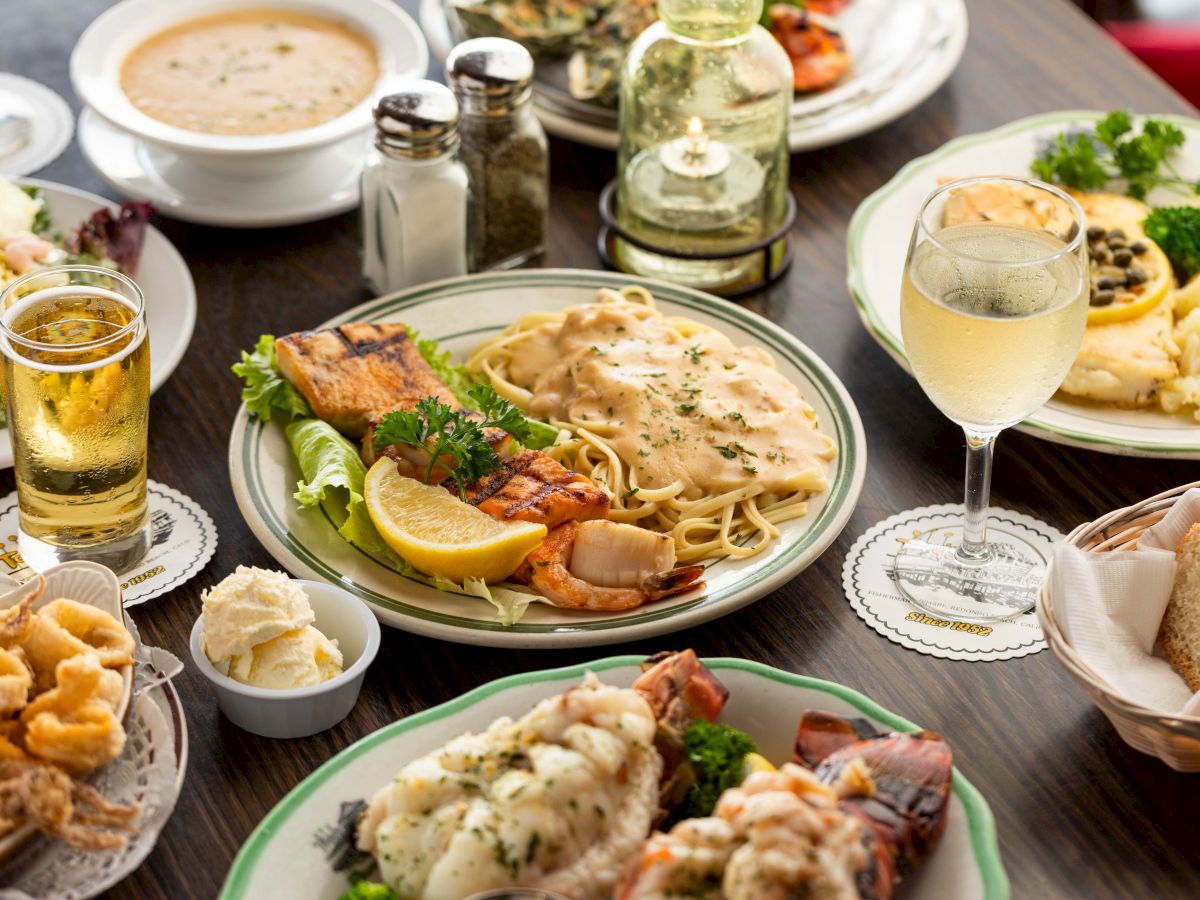 A spread of seafood pasta with shrimp and lemon, salad, garlic sauce, bread and rolls, plus glasses of beer and sparkling wine.