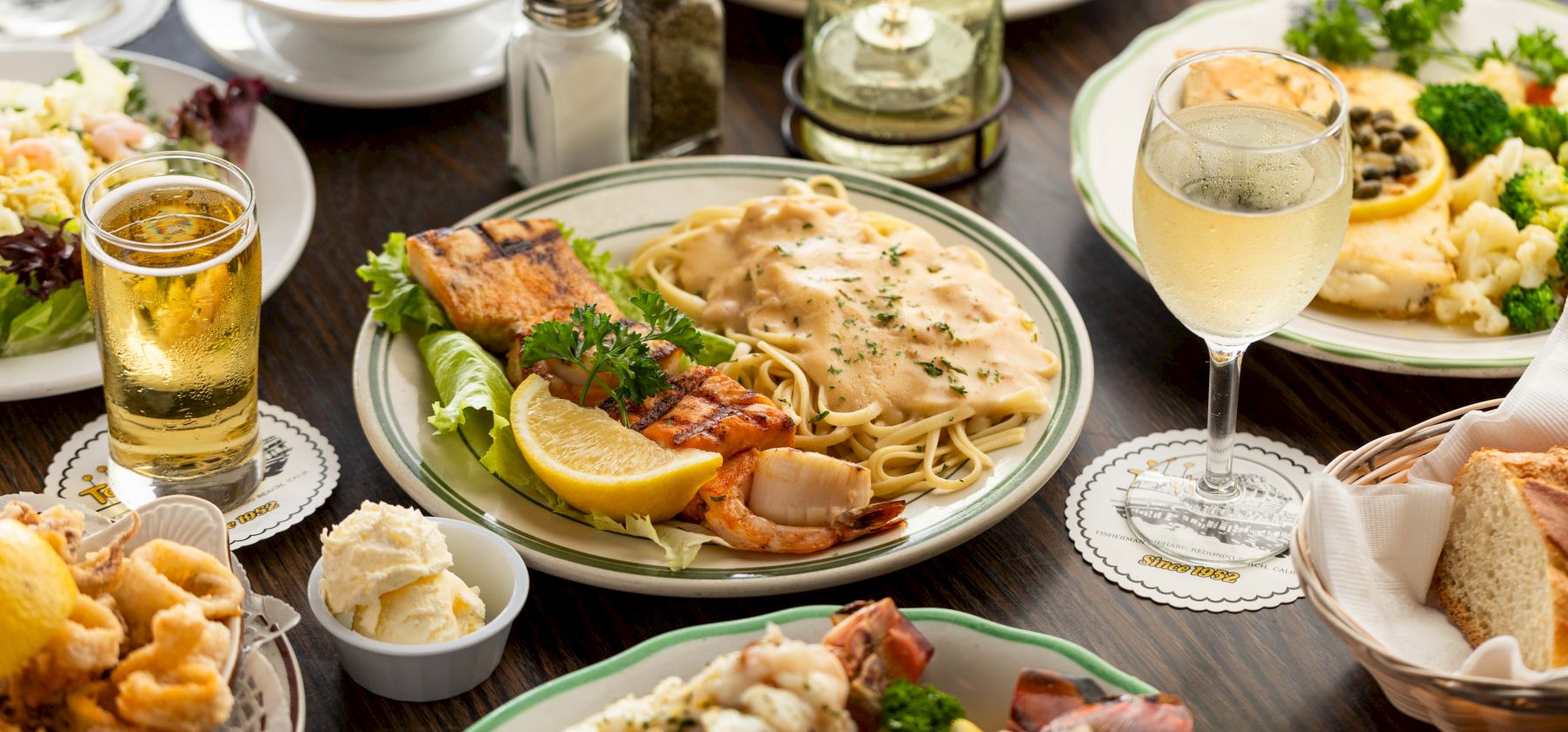 A spread of seafood pasta with shrimp and lemon, salad, garlic sauce, bread and rolls, plus glasses of beer and sparkling wine.