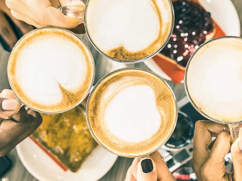 A cheerful group cheers with latte art-topped coffees, toasting friends over a shared brunch.