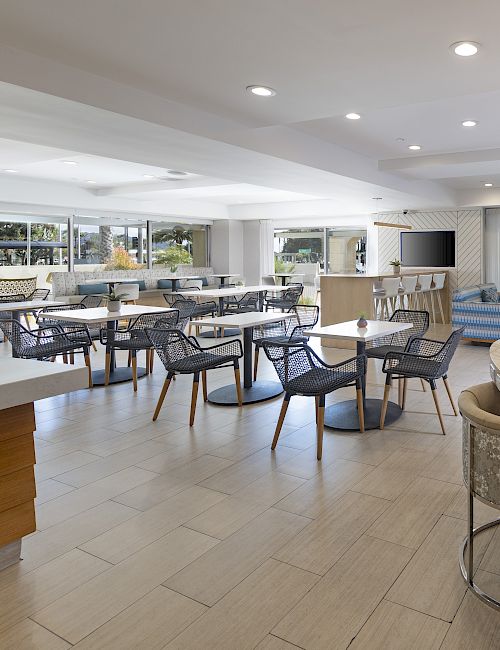 A bright modern patio lounge with wicker tables and chairs, a long wooden bar counter, and outdoor seating under ceiling lights.