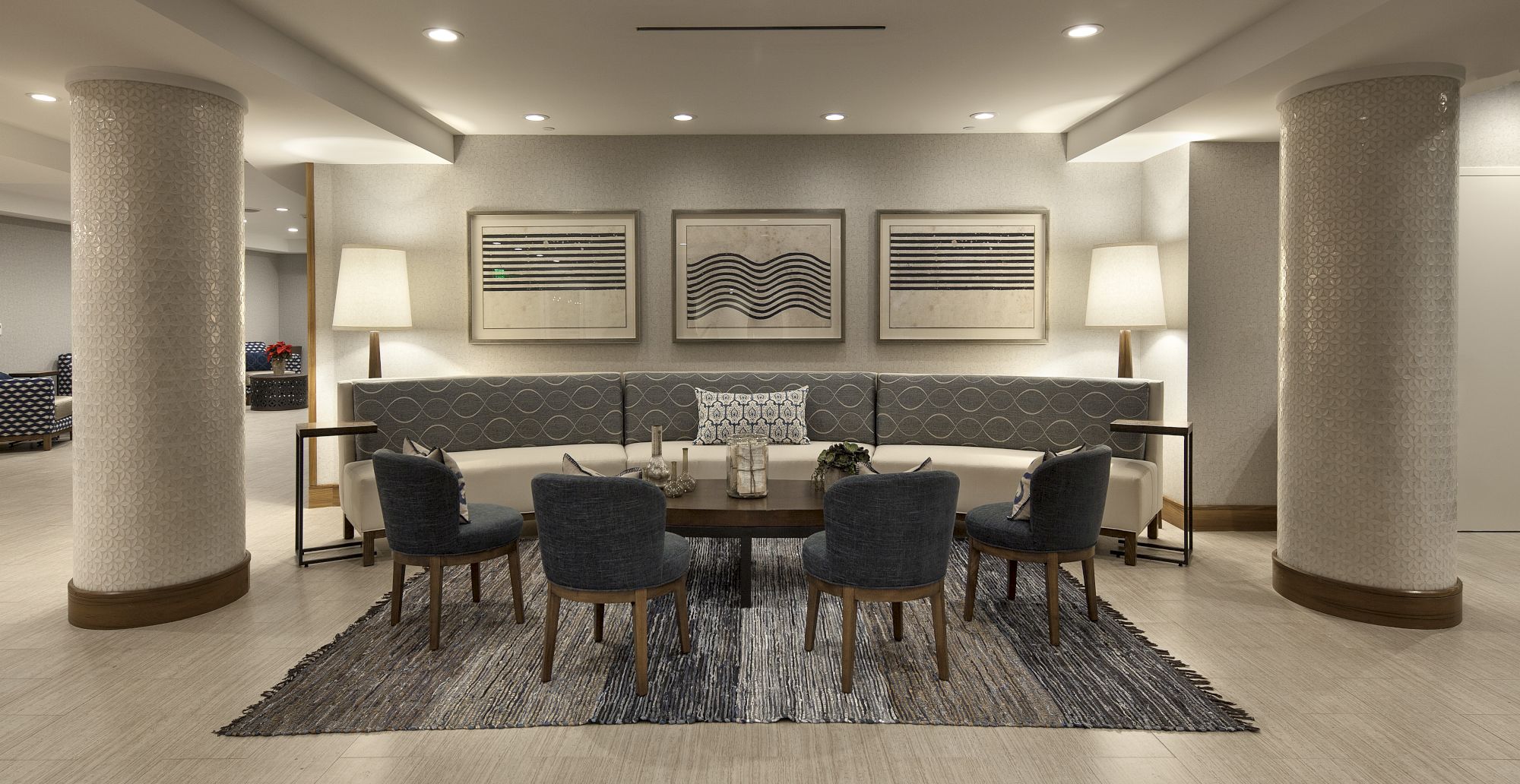 A modern lounge with a central round table, eight chairs, soft lighting, framed art, and a patterned rug on a light, airy space.