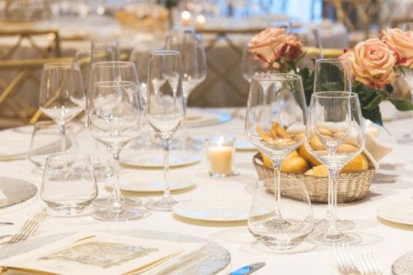 A formal dining table setup with empty wine glasses, plates, cutlery, candle, and a bouquet of peach roses in the background.