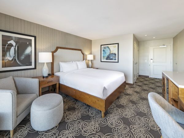 A modern hotel room with a king bed, bedside lamps, framed art, a sofa, patterned carpet, and a vanity desk.