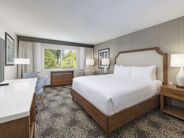 A modern hotel bedroom with a large bed, nightstands, lamps, framed art, and a window showing palm trees outside.