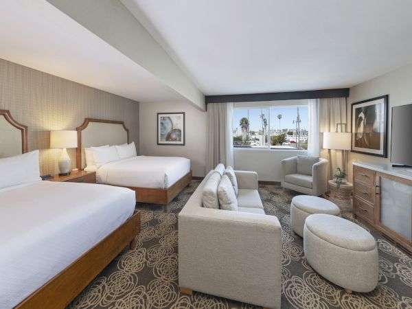 A hotel room with two beds, lamps, a sofa chair, a TV/desk area, and a large window view outside.