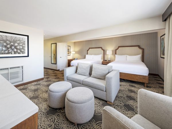 A bright hotel room with two beds, a gray sofa, patterned carpet, framed art, and bedside lamps.