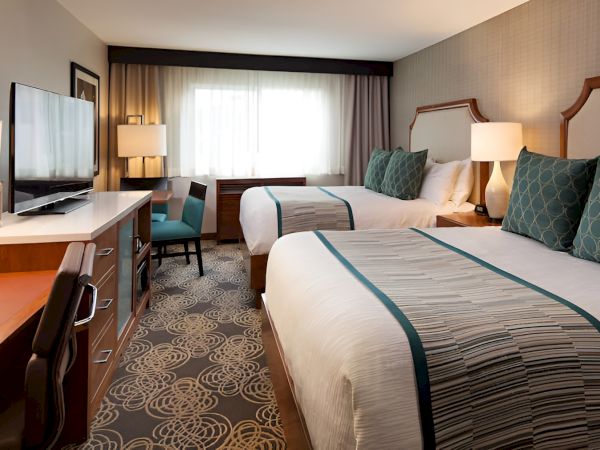 A modern hotel room with two queen beds, a desk, flat-screen TV, desk chair, nightstands, lamps, and a window letting in natural light.