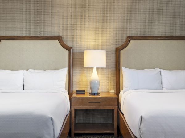 Two neatly made beds in a hotel room, with a central nightstand and warm table lamps against a textured wall.