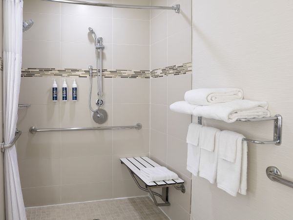 A clean, accessible shower with grab bars, showerhead, tiled walls, toiletries bottles, and neatly folded white towels.