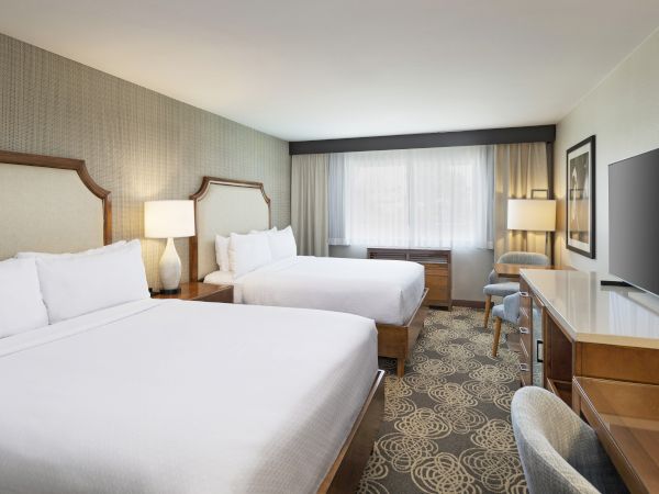 A modern hotel room with two neatly made beds, bedside lamps, curtains, and a flat-screen TV on the dresser.