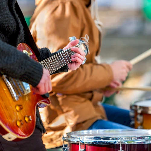Guitar and drums being played in a band scene, close-up of hands on strings and sticks, outdoors, casual jam session.