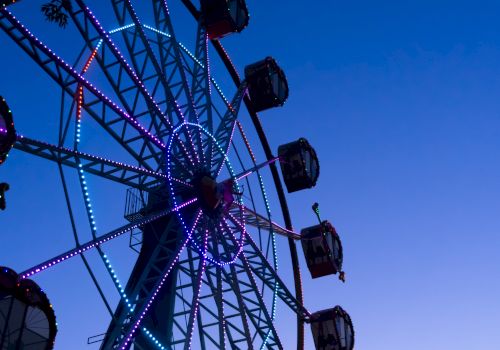 A colorful Ferris wheel lit with purple and blue lights against a twilight sky, with passenger gondolas along the rim.