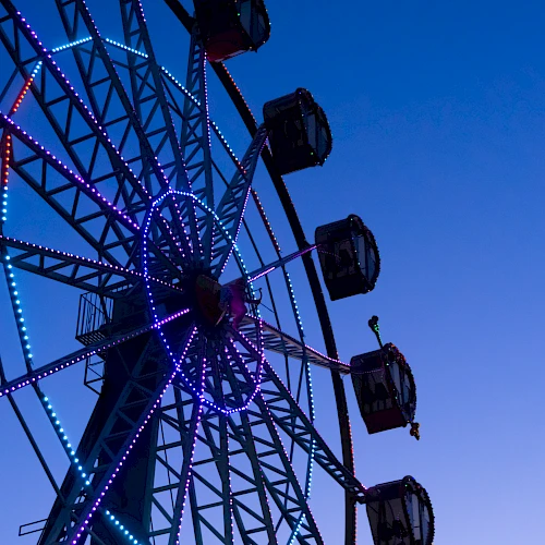 A colorful Ferris wheel lit with purple and blue lights against a twilight sky, with passenger gondolas along the rim.