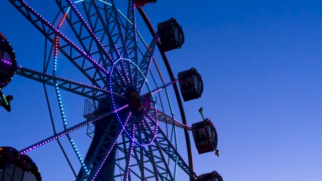 A colorful Ferris wheel lit with purple and blue lights against a twilight sky, with passenger gondolas along the rim.