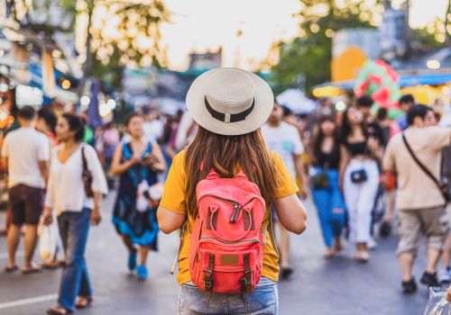 A person with a pink backpack stands in a busy outdoor market or street festival, wearing a tan hat and yellow shirt, facing a crowd.