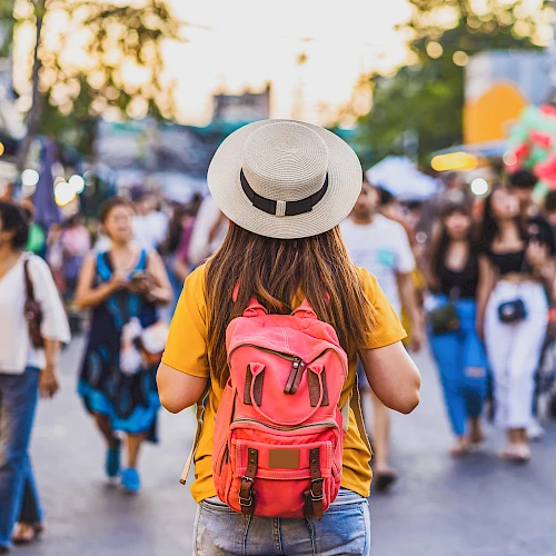 A person with a pink backpack stands in a busy outdoor market or street festival, wearing a tan hat and yellow shirt, facing a crowd.