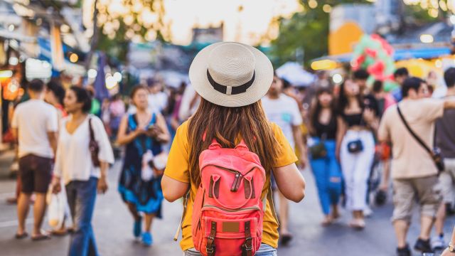 A person with a pink backpack stands in a busy outdoor market or street festival, wearing a tan hat and yellow shirt, facing a crowd.
