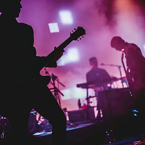 A rock band performing on stage with silhouetted guitarists, drummer, and vibrant purple lighting.