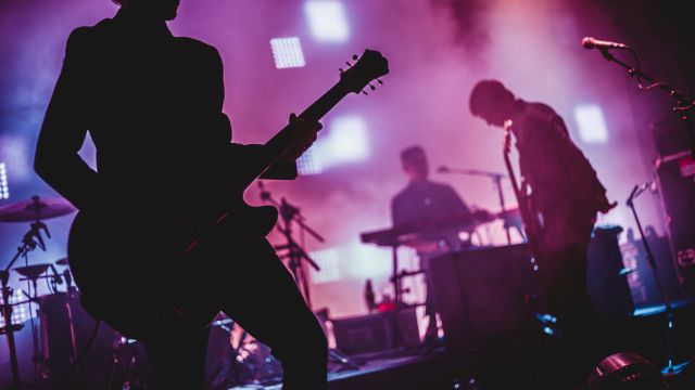 A rock band performing on stage with silhouetted guitarists, drummer, and vibrant purple lighting.