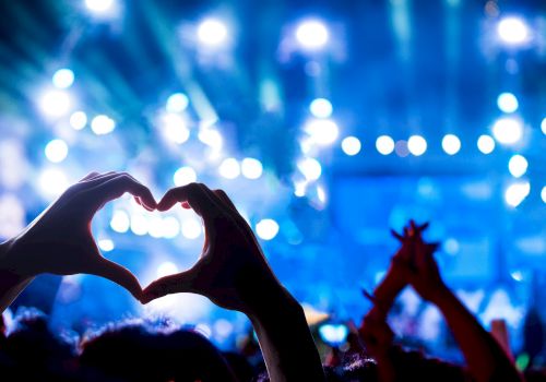 A lively concert scene with hands forming a heart shape in the foreground, blue stage lights, and a cheering crowd.