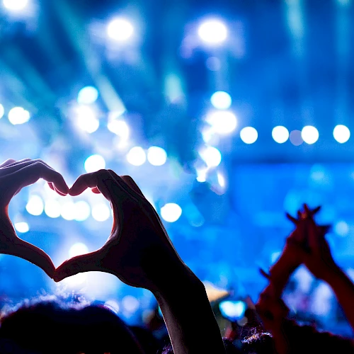 A lively concert scene with hands forming a heart shape in the foreground, blue stage lights, and a cheering crowd.