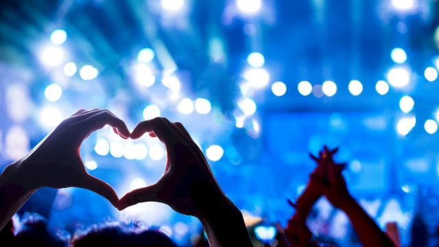 A lively concert scene with hands forming a heart shape in the foreground, blue stage lights, and a cheering crowd.