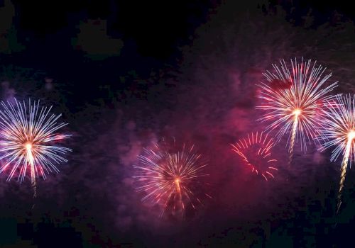 Colorful red, white, and pink fireworks burst across a dark sky with smoky clouds.
