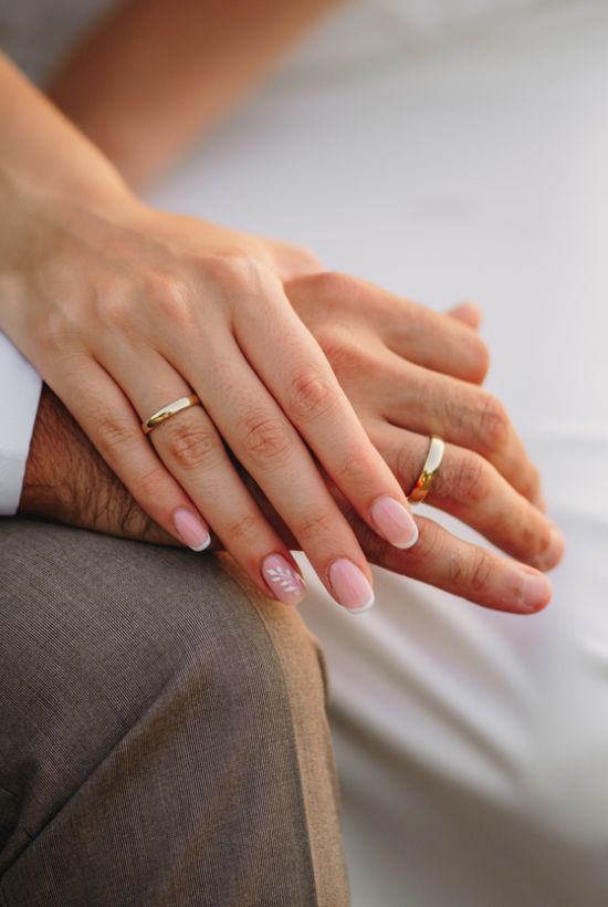 A bride and groom hold hands, showing wedding rings and manicured nails in warm daylight.