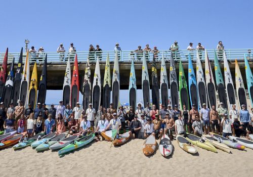 A group of people pose on a sunny beach behind a row of colorful racing kayaks and flags atop a pier, together in one team photo.