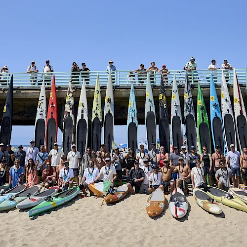 A group of people pose on a sunny beach behind a row of colorful racing kayaks and flags atop a pier, together in one team photo.