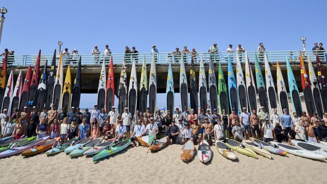 A group of people pose on a sunny beach behind a row of colorful racing kayaks and flags atop a pier, together in one team photo.