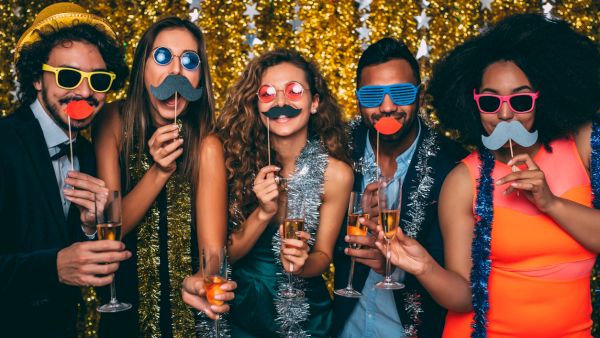 A group of friends at a party in front of golden tinsel, holding champagne and wearing funny sunglasses and mustaches.